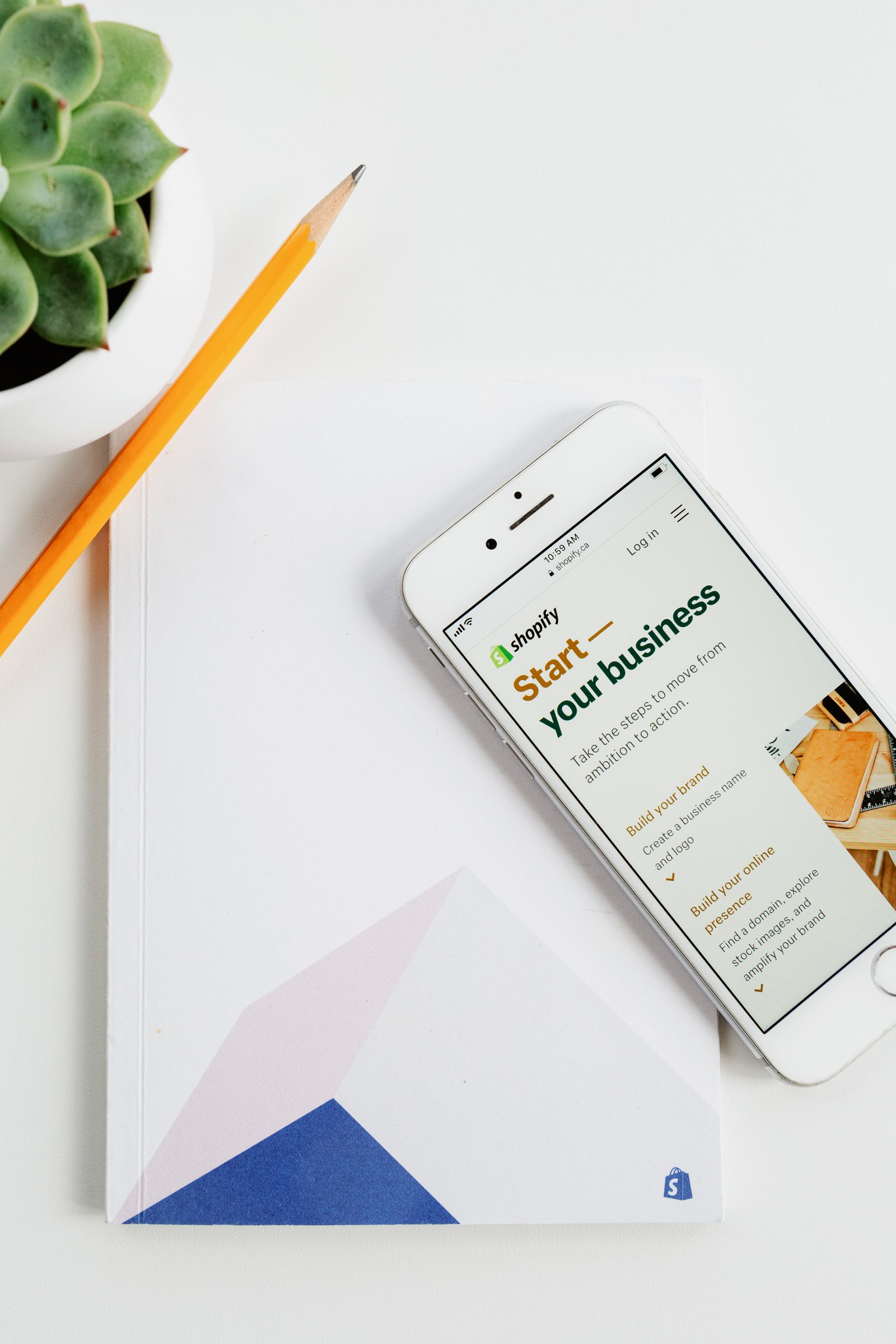 A phone, pencil, notepad and plant on a white table. Displayed on the phone is the Shopify website.